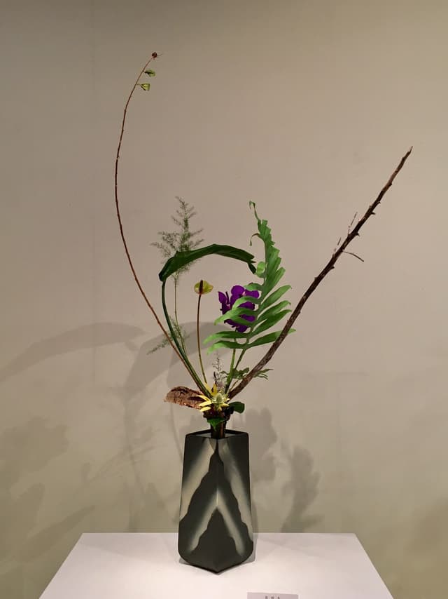 Classical Ikenobō ikebana with a curved branch and purple blossoms in a dark angular vase
